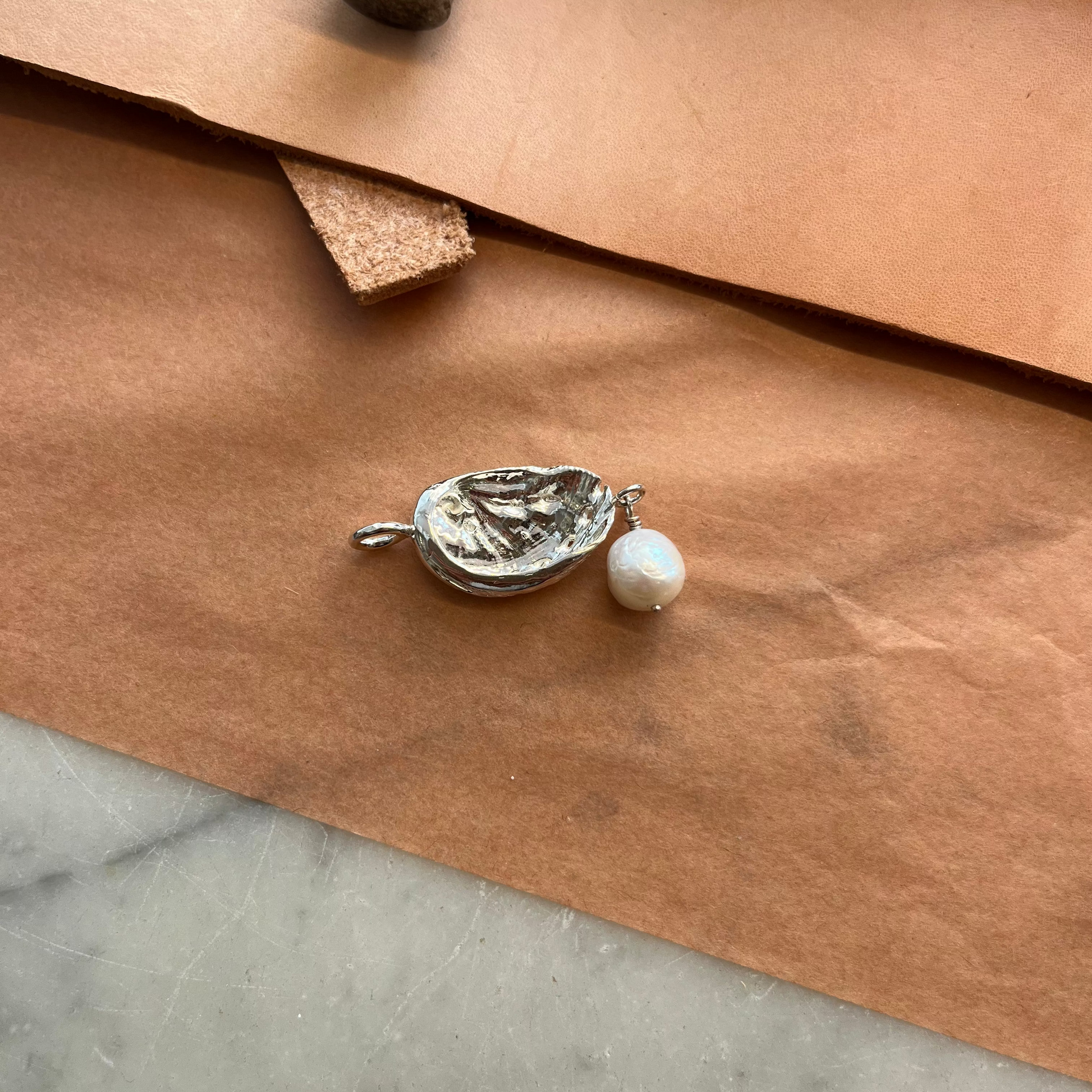 A silver shell-shaped pendant with a small white pearl charm resting on sheets of brown paper, with a hammer and leather tool partially visible in the background.