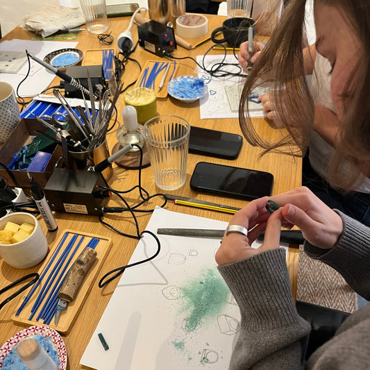 A top-down view of a workshop with a craft table with multiple people' hands and arms visible, working on various projects including drawing on white paper, using tools, and holding small objects. A cup of dark tea and cookies on a plate are also visible on the wooden table.